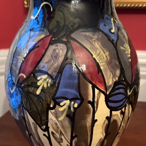 May include: A ceramic vase with a black background and a floral design. The flowers are painted in shades of blue, red, and yellow. The vase has a rounded body and a flared lip.