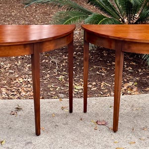 May include: Two matching, semi-circular wooden tables with a rich, warm brown finish. Each table has four slender, tapered legs. The tables are positioned side-by-side, showcasing their similar design and craftsmanship. The tables are approximately 30 inches wide.