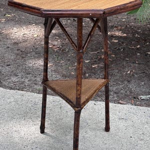 May include: A small, octagonal side table with a woven top and a triangular shelf. The table is made of dark brown bamboo and stands approximately 28 inches tall.