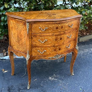 May include: A three-drawer chest with a curved front and intricate marquetry details. The chest is made of wood with a rich brown finish and features brass hardware. The chest is sitting on a black surface.