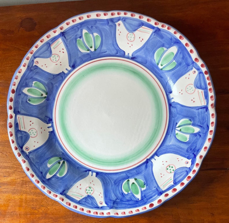 K&ouml;nnte beinhalten: Ein gro&szlig;er, runder Teller aus Keramik mit einem blauen und wei&szlig;en Design. Der Teller hat einen Rand mit roten Punkten und ein sich wiederholendes Muster aus stilisierten V&ouml;geln und gr&uuml;nen Bl&auml;ttern.