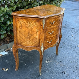 May include: A curved, wooden chest of drawers with three drawers. The chest is made of a light brown wood with intricate marquetry details. The drawers have brass handles and the chest has brass feet.