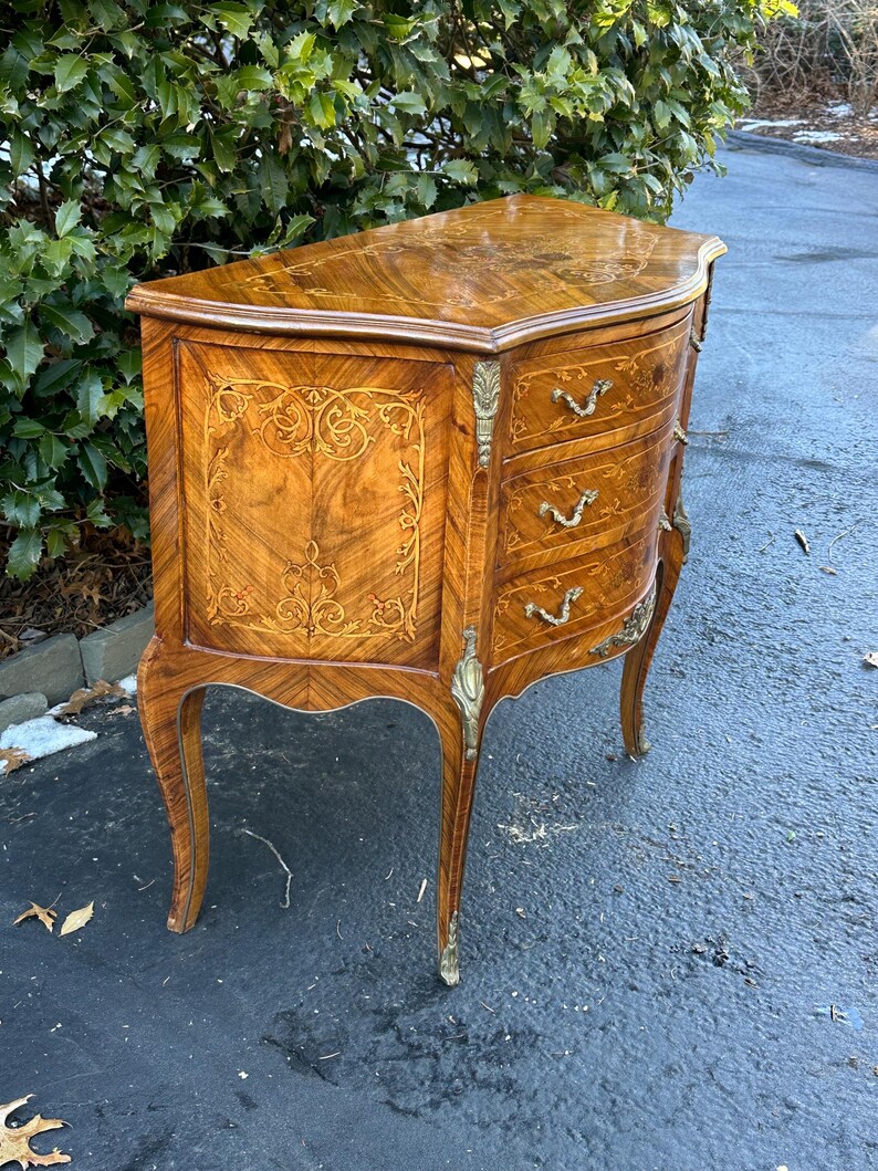 May include: A curved, wooden chest of drawers with three drawers. The chest is made of a light brown wood with intricate marquetry details. The drawers have brass handles and the chest has brass feet.