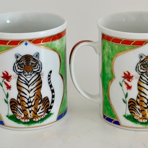May include: Two white ceramic mugs with a tiger design. Each mug features a seated tiger with orange and black stripes, set against a white background with a green and red border. The mugs have white handles and are suitable for hot beverages.