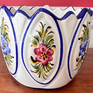 May include: White ceramic planter with blue and yellow floral designs. The planter has a scalloped rim and is decorated with blue and yellow flowers and green leaves.