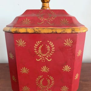 May include: A hexagonal red canister with a lid featuring a gold knob. The body and lid are decorated with gold-colored laurel wreath motifs and small star-like patterns. The canister rests on four small, mottled yellow and brown feet.