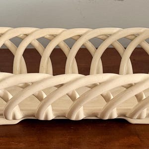 May include: Cream-colored, oval-shaped ceramic basket with a woven design. The basket features a series of arched, intertwined supports, creating an openwork pattern. The basket has a handle on each end and sits on a flat base.