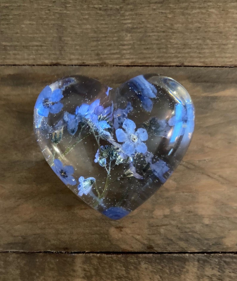 Puede incluir: Un pisapapeles de resina transparente en forma de coraz&oacute;n con flores azules de nomeolvides prensadas en su interior.