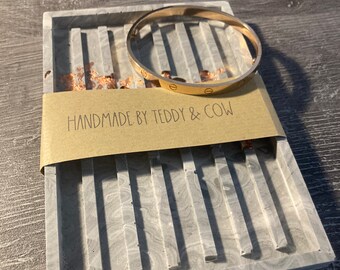 May include: A gray concrete tray with a gold-toned bangle on top. The tray has a marbled pattern and the text "HANDMADE BY TEDDY & COW" is printed on a brown paper label.