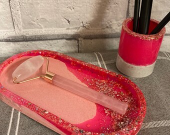 May include: A pink and white oval tray with glitter accents, featuring a pink rose quartz facial roller with a gold handle.