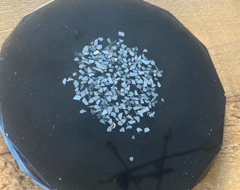 May include: A black round coaster with a circular design of small, black and silver glitter pieces in the center.