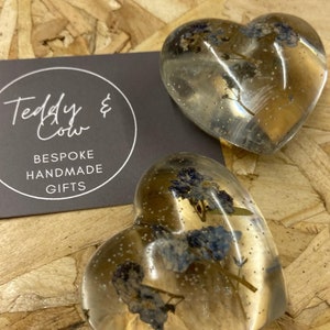 Puede incluir: Dos adornos de resina transparentes en forma de coraz&oacute;n con flores azules secas prensadas en su interior. Los adornos est&aacute;n sobre un fondo de madera. El texto "Teddy & Cow Bespoke Handmade Gifts" est&aacute; en una tarjeta negra detr&aacute;s de los adornos.