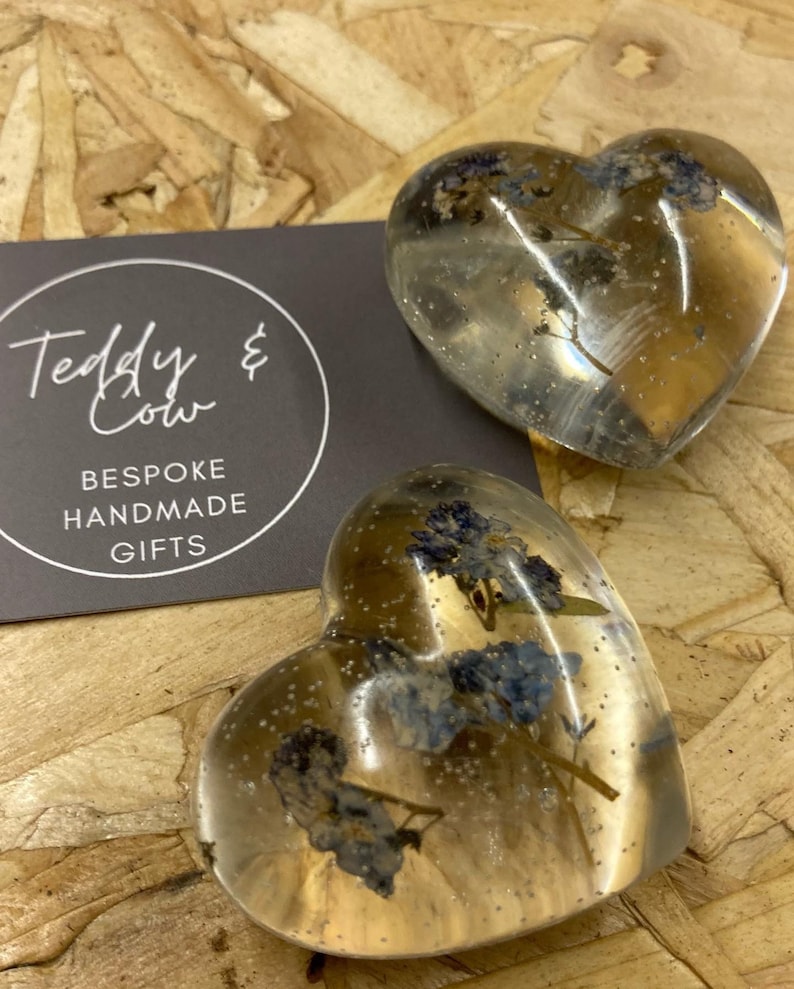 Puede incluir: Dos adornos de resina transparentes en forma de coraz&oacute;n con flores azules secas prensadas en su interior. Los adornos est&aacute;n sobre un fondo de madera. El texto "Teddy & Cow Bespoke Handmade Gifts" est&aacute; en una tarjeta negra detr&aacute;s de los adornos.