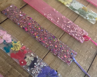 Puede incluir: Un conjunto de seis marcapáginas rosas y transparentes con flores prensadas y purpurina. Los marcapáginas tienen un borla en la parte inferior.