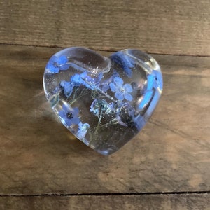 Puede incluir: Un pisapapeles de resina transparente en forma de coraz&oacute;n con flores azules de nomeolvides prensadas en su interior.