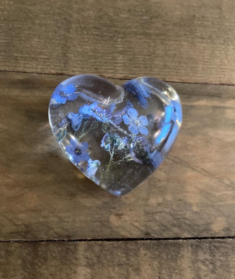 Puede incluir: Un pisapapeles de resina transparente en forma de coraz&oacute;n con flores azules de nomeolvides prensadas en su interior.