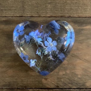 Puede incluir: Un pisapapeles de resina transparente en forma de coraz&oacute;n con flores azules de nomeolvides prensadas en su interior.