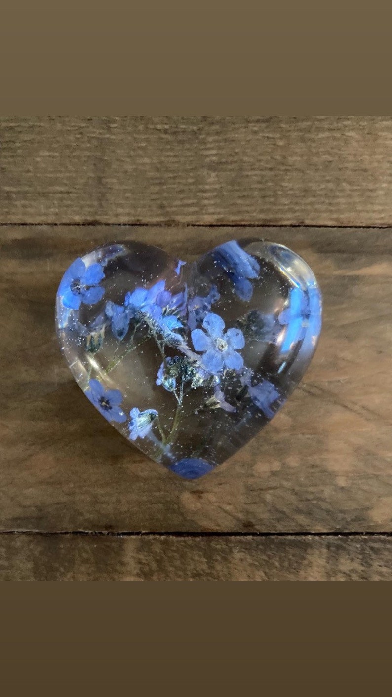 Puede incluir: Un pisapapeles de resina transparente en forma de coraz&oacute;n con flores azules de nomeolvides prensadas en su interior.