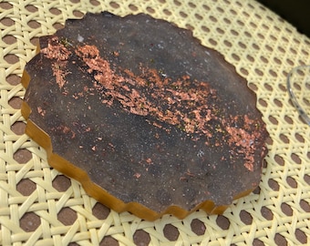 Puede incluir: Un posavasos redondo de resina negra y dorada con escamas de cobre incrustadas en la resina. El posavasos tiene un borde festoneado.