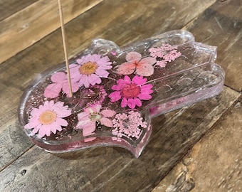 Puede incluir: Un porta incienso de resina transparente con forma de mano de Hamsa con flores prensadas rosas en su interior. Un palo de incienso de madera está en el porta incienso.