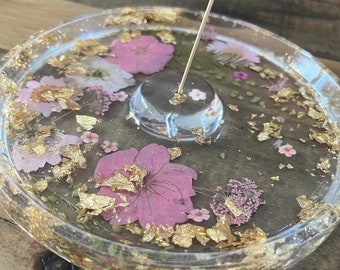 May include: A clear glass incense holder with a raised platform for the incense stick. The holder is decorated with pressed pink and white flowers and gold leaf flakes.