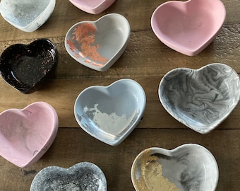 May include: A collection of ten heart-shaped trinket dishes in various colors and patterns. The dishes are made of concrete and feature marbled, glitter, and metallic finishes.