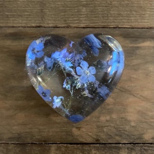 Puede incluir: Un pisapapeles de resina en forma de coraz&oacute;n con flores de nomeolvides azules prensadas en su interior.