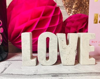 May include: White concrete letters spelling out the word "LOVE" on a gray and white textured surface. The letters are in focus and the background is blurred. There are pink honeycomb decorations and a pink card with gold lettering that says "All I Need Is... But I'll Accept Chocolate" in the background.