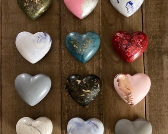 May include: Twelve heart-shaped decorations in various colors and patterns, including marbled, speckled, and solid colors. The hearts are arranged in three rows of four, on a wooden background.