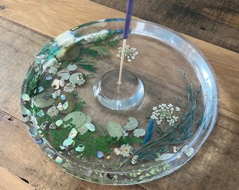 May include: A clear glass incense holder with a round base and a raised center for holding incense sticks. The holder is decorated with pressed flowers and leaves, including white flowers, green leaves, and blue accents.