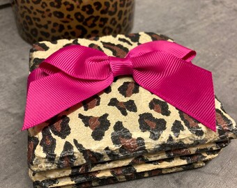 May include: A set of three square coasters with a leopard print design. The coasters are stacked and tied together with a bright pink bow.