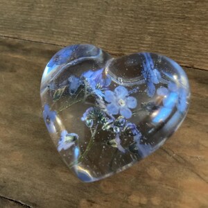 Puede incluir: Un pisapapeles transparente en forma de coraz&oacute;n con flores azules de nomeolvides prensadas en su interior.