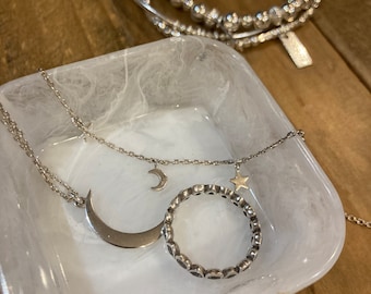 May include: A silver necklace with a crescent moon pendant and a star charm, a silver ring with a circle of small hearts, and a silver bracelet with a circle of small beads, all displayed on a white marble dish.