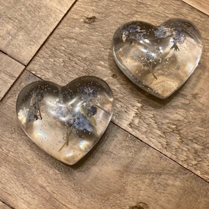 Puede incluir: Dos adornos de resina transparentes en forma de coraz&oacute;n con flores azules secas en su interior. Los adornos est&aacute;n sobre una superficie de madera.