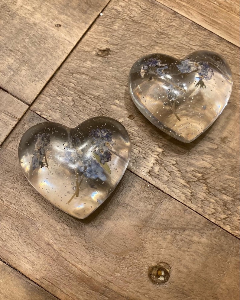 Puede incluir: Dos adornos de resina transparentes en forma de coraz&oacute;n con flores azules secas en su interior. Los adornos est&aacute;n sobre una superficie de madera.