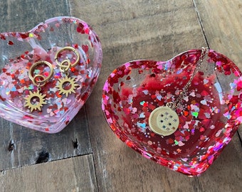 May include: Two heart-shaped resin trinket dishes with glitter and confetti. One dish is clear with pink and white glitter and features gold earrings. The other dish is red with pink and white glitter and features a gold necklace with a star pendant.