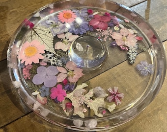 May include: A clear glass coaster with a round, raised center and a decorative border of pressed flowers. The flowers are pink, purple, and white, and are arranged in a random pattern. The coaster is perfect for protecting surfaces from hot drinks or other items.
