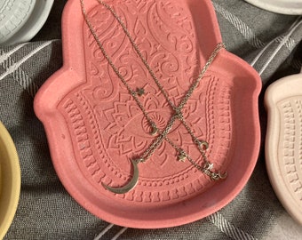 May include: A pink hamsa hand tray with a decorative design and three silver necklaces with star and crescent moon charms.