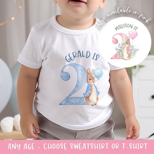 Può includere: T-shirt bianca con un'illustrazione ad acquerello di Peter Coniglio che tiene un palloncino blu, con la scritta "GERALD IS 2". Un'immagine più piccola mostra un design simile in rosa con la scritta "MADISON IS". Il testo "ANY AGE - CHOOSE SWEATSHIRT OR T-SHIRT" è in basso.
