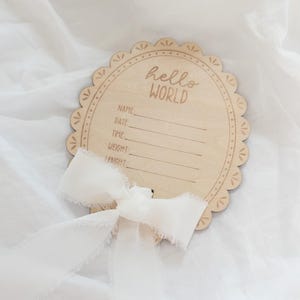Pode incluir: Uma placa de madeira redonda com borda recortada apresentando o texto gravado hello WORLD e linhas para Name, Date, Time, Weight e Length. Uma fita branca desfiada está amarrada em um laço na parte inferior da placa de madeira clara.