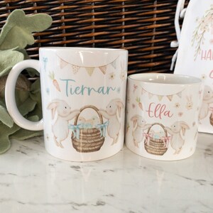 K&ouml;nnte beinhalten: Wei&szlig;er Keramikbecher und Tasse mit Ostermotiv. Die Designs zeigen Hasen, Osterk&ouml;rbe und die Namen "Tiernan" und "Ella". Ein passender Kordelzugbeutel mit "Happy Easter Sophie" ist ebenfalls zu sehen.