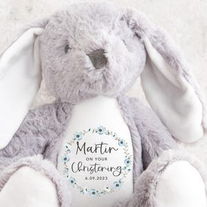 May include: A plush gray bunny rabbit with long ears and white accents on the paws and inner ears. The bunny has a white patch on its chest with the text "Martin on your Christening 6.09.2023" surrounded by a blue floral wreath.