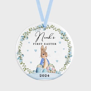 Puede incluir: Adorno de Pascua redondo y blanco con una cinta azul claro. Presenta una ilustración en acuarela de un conejito con chaqueta azul, rodeado de huevos de Pascua y una corona floral. El texto dice "Noah's First Easter 2024."