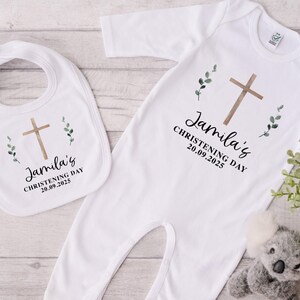 Peut inclure: Ensemble de vêtements blancs pour bébé, comprenant un bavoir et une combinaison, ornés d'une croix marron, de verdure et de l'inscription "Jamila's Christening Day 20.09.2025". Un petit koala en peluche est visible en arrière-plan.