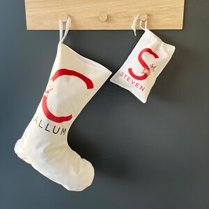 May include: A white Christmas stocking featuring a large red letter C with a small Santa graphic and the name CALLUM in black text, alongside a smaller white gift bag featuring a large red letter S with a reindeer graphic and the name STEVEN.
