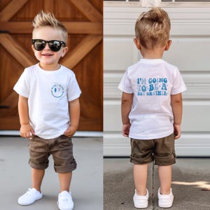 以下が含まれることがあります： 笑顔と稲妻のデザインが施された白いTシャツ、茶色のショートパンツ、白いスニーカー、サングラスを着用した幼い子供。Tシャツの背面には「I'M GOING TO BE A BIG BROTHER」と書かれています。