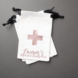 Puede incluir: Bolsas blancas con cordón y una cruz de oro rosa y el texto "Lauren's Bachelorette". Las bolsas tienen lazos de cinta negra. Las bolsas están apiladas una encima de otra.