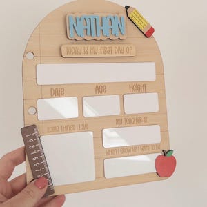 Op de afbeelding: Een houten bord voor de eerste schooldag in de vorm van een notitieblok met de naam Nathan in blauw, een geel potlood en een rode appel. Het heeft witte uitwisbare vakken voor datum, leeftijd, lengte, interesses, leraar en doelen, met een liniaal van 20 cm.