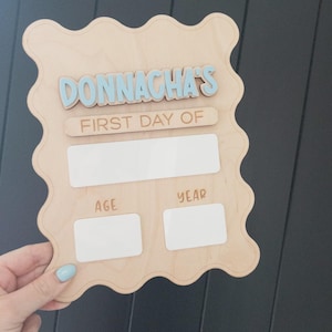 Könnte beinhalten: Ein helles Holzschild mit gewelltem Rand und dem Schriftzug DONNACHA'S FIRST DAY OF in erhabenen Buchstaben. Das Schild enthält ein großes weißes rechteckiges Feld sowie zwei kleinere weiße rechteckige Felder mit der Beschriftung AGE und YEAR.