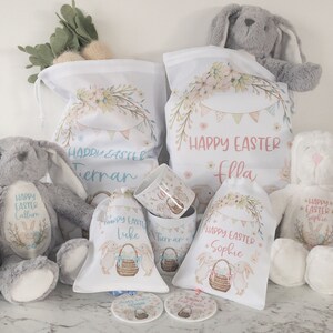 K&ouml;nnte beinhalten: Eine Auswahl an Ostergeschenken, darunter Pl&uuml;schhasen, Kordelzugbeutel und Tassen. Die Beutel und Tassen sind mit Blumen- und Wimpeldesigns, dem Text "Frohe Ostern" und Namen verziert. Die Hasen sind grau und wei&szlig;.